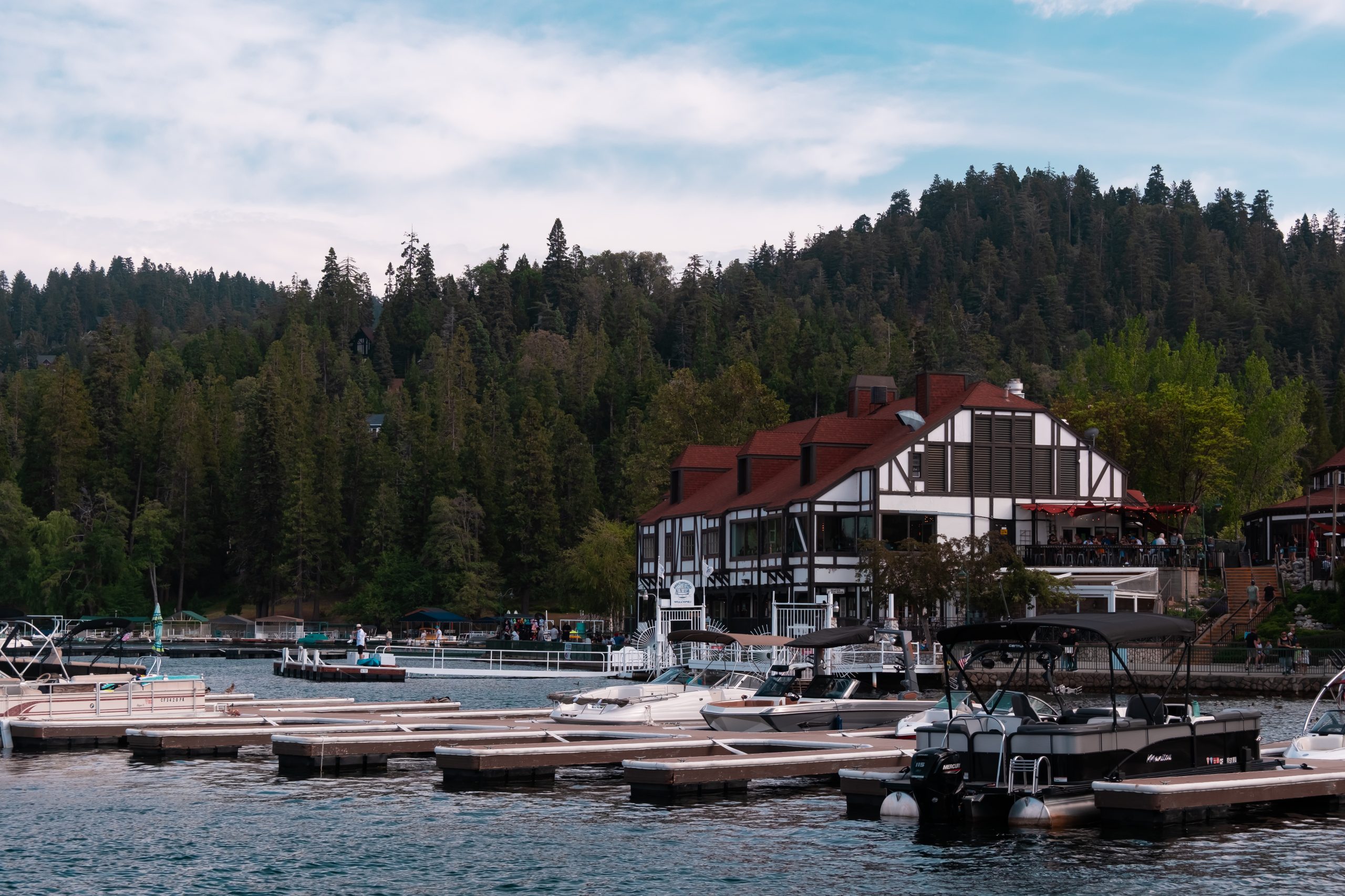 View of Lake Arrowhead with moored boats and the Lake Arrowhead Village visible along the forested shoreline under a cloudy sky.