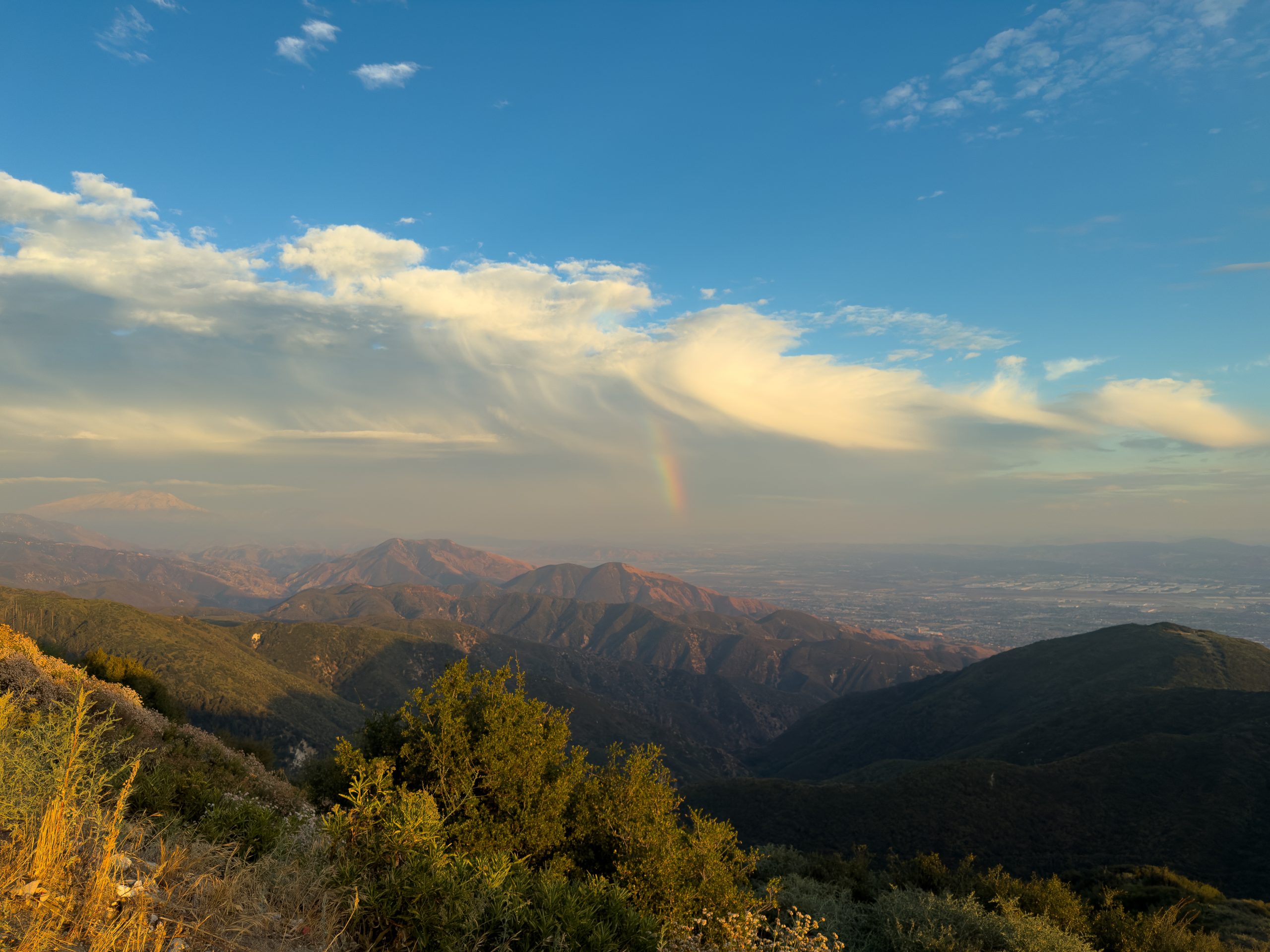 View from a high viewpoint overlooking the San Bernardino Valley with mountains under a blue sky, featuring a faint rainbow and the sun setting in the distance.