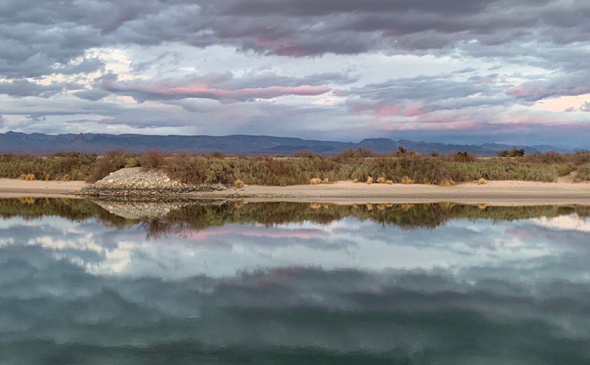 A view of the Colorado River under some colorful clouds with mountains in the background.