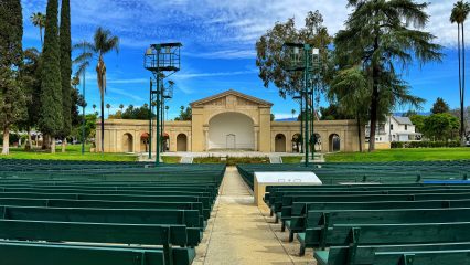 View of the Redlands Bowl with green benches and a stage, surrounded by trees under a blue sky with clouds.