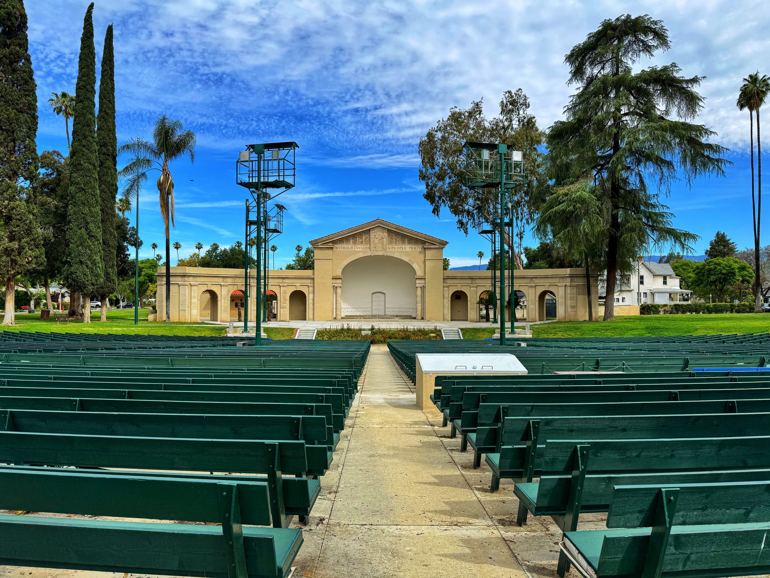 View of the Redlands Bowl with green benches and a stage, surrounded by trees under a blue sky with clouds.