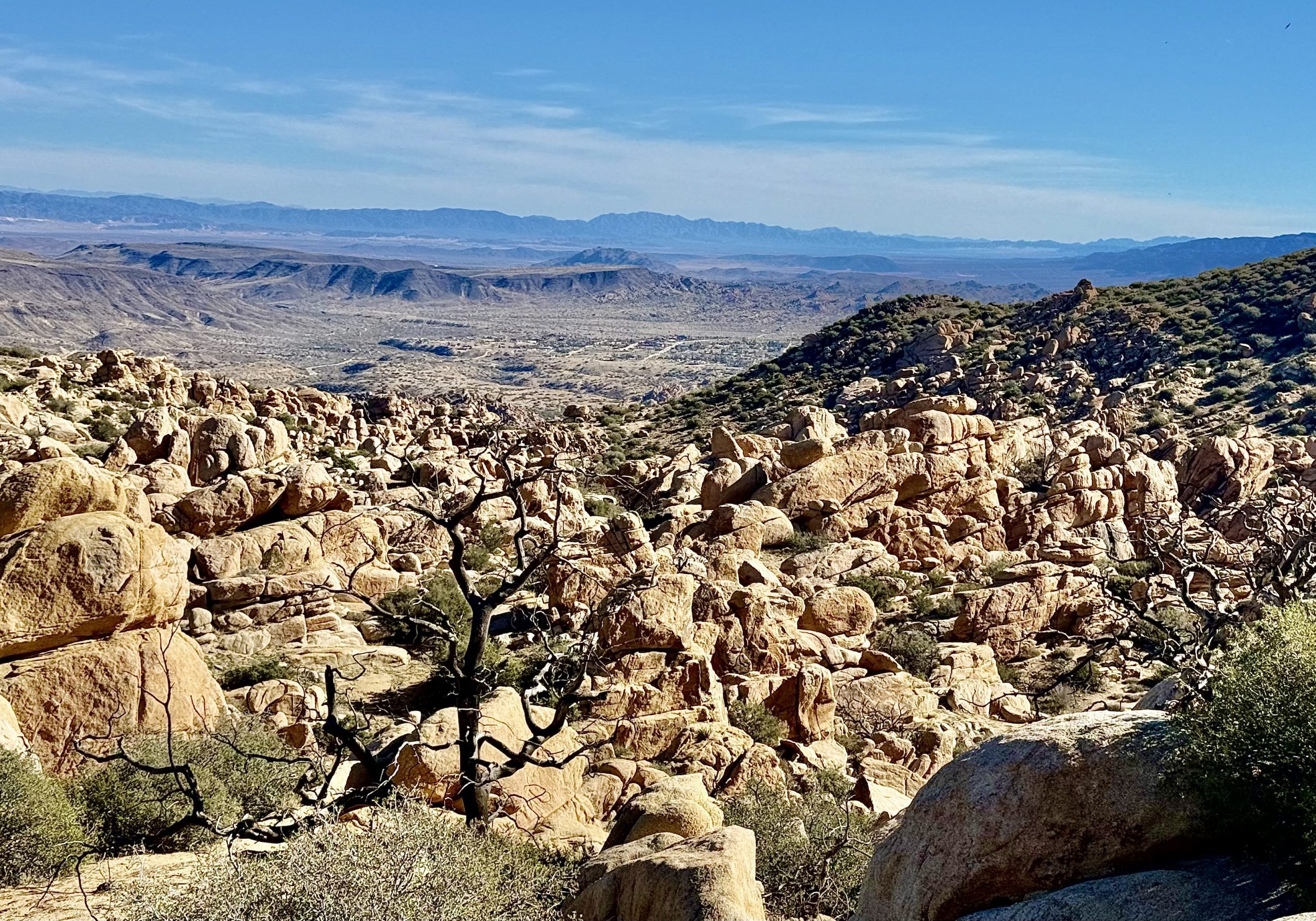 A panoramic view of a rugged landscape filled with large boulders and scattered desert vegetation. Sunny skies and distant mountain ranges complete the scene.