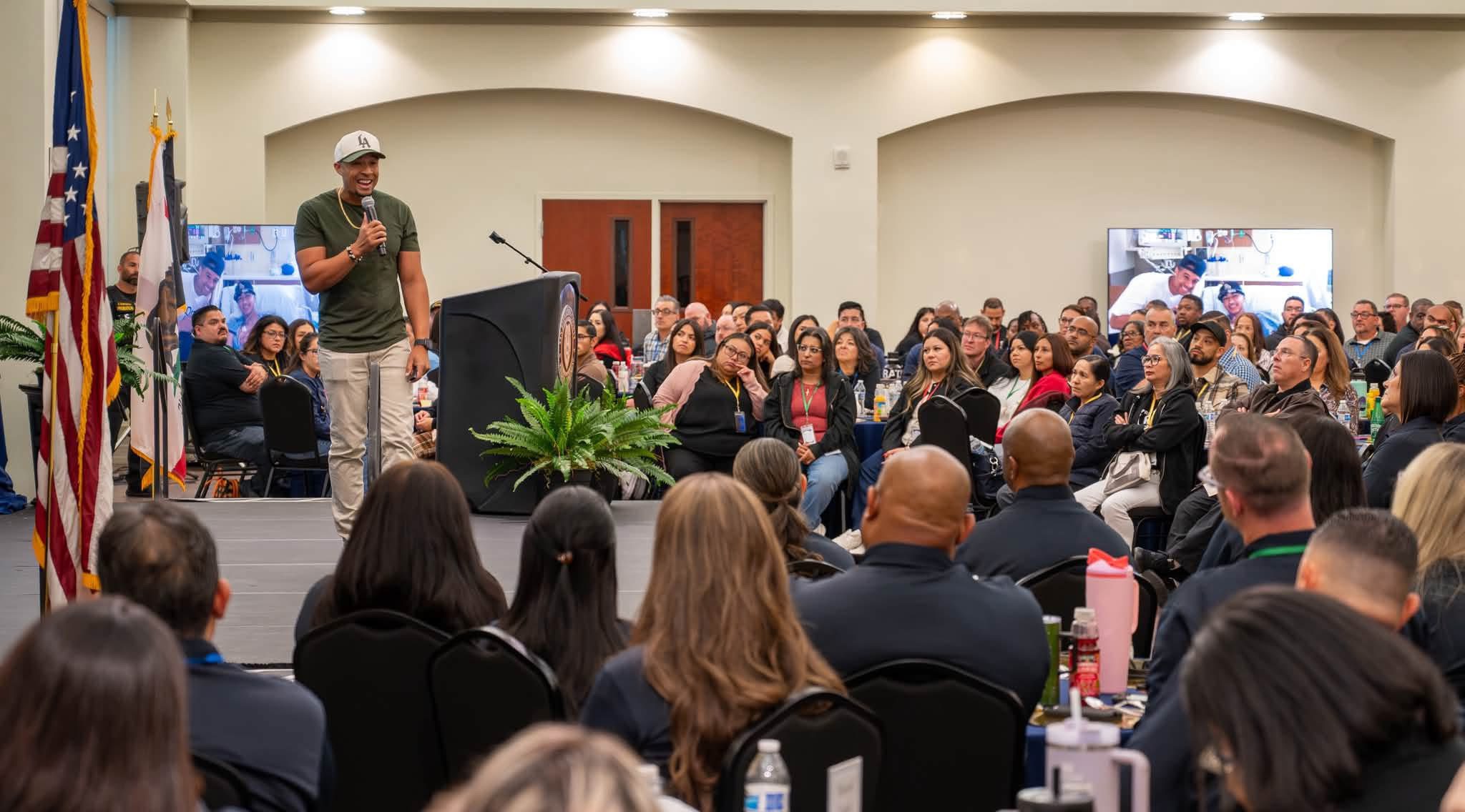A person is speaking on a stage to a large audience seated in a conference room