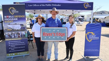 San Bernardino County "Law Offices of the Public Defender" canopy and three people holding "The River’s Edge Ranch” sign at mobile unit event.