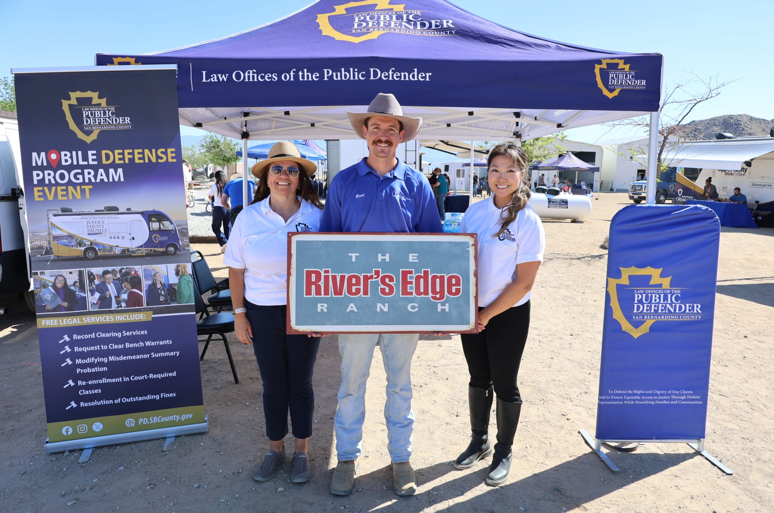 San Bernardino County "Law Offices of the Public Defender" canopy and three people holding "The River’s Edge Ranch” sign at mobile unit event.
