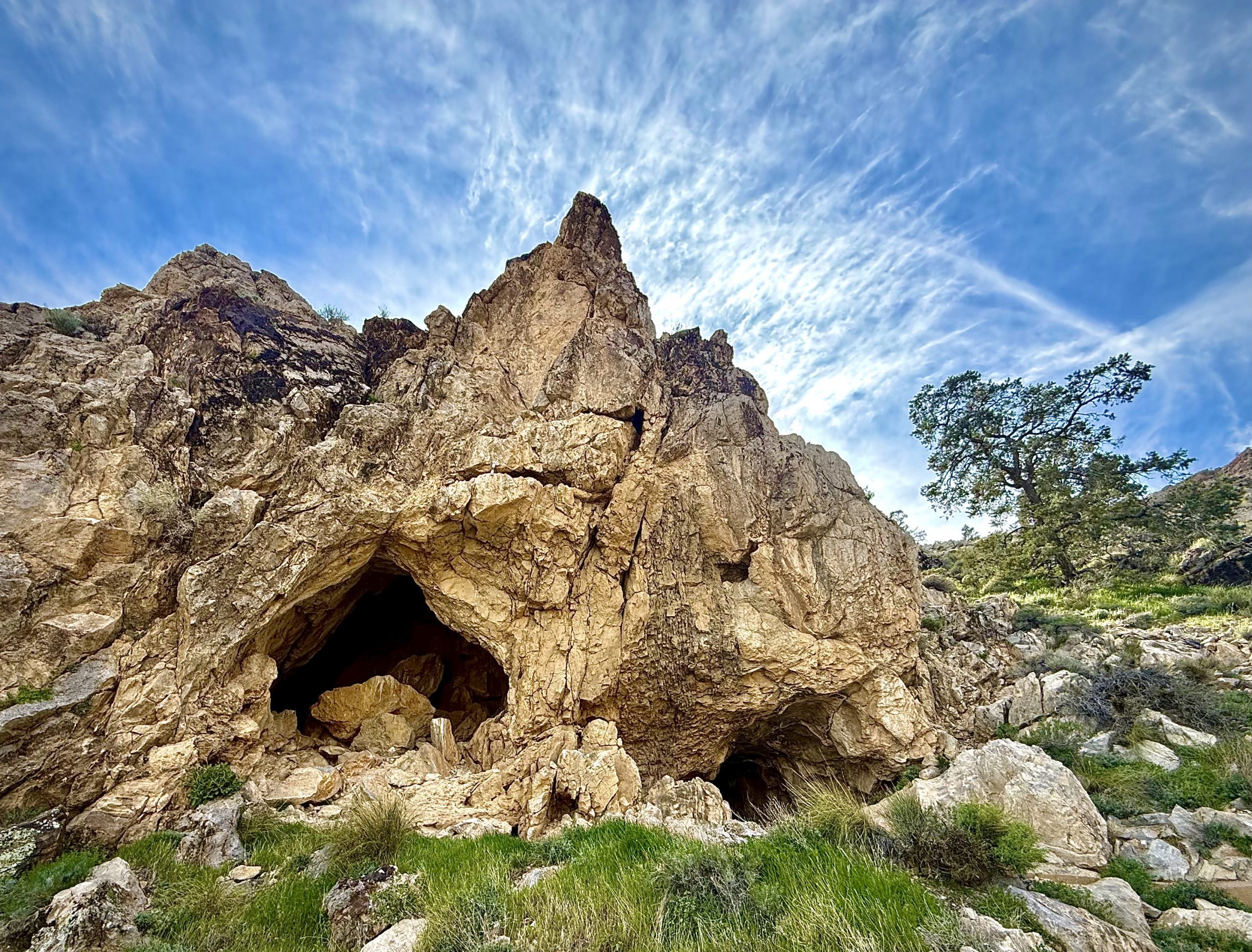 A rocky hill with two cave openings in the foreground. The sky is clear with a few wispy clouds, and a tree stands to the right.