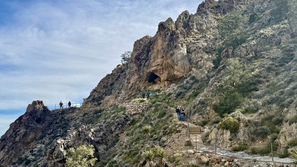 A rocky hillside with a trail leads to a cave entrance. Several people are walking along the path.