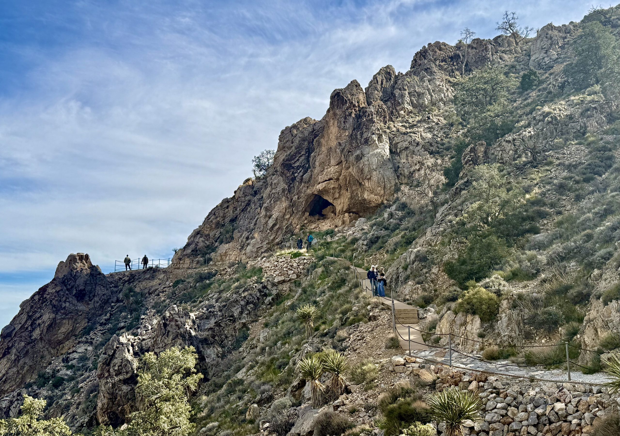 A rocky hillside with a trail leads to a cave entrance. Several people are walking along the path.