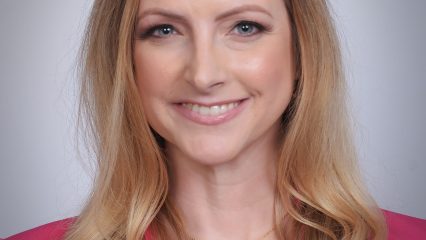 Smiling woman with long blonde hair wearing a pink blazer and heart-shaped necklace. Neutral background. Confident and friendly expression.