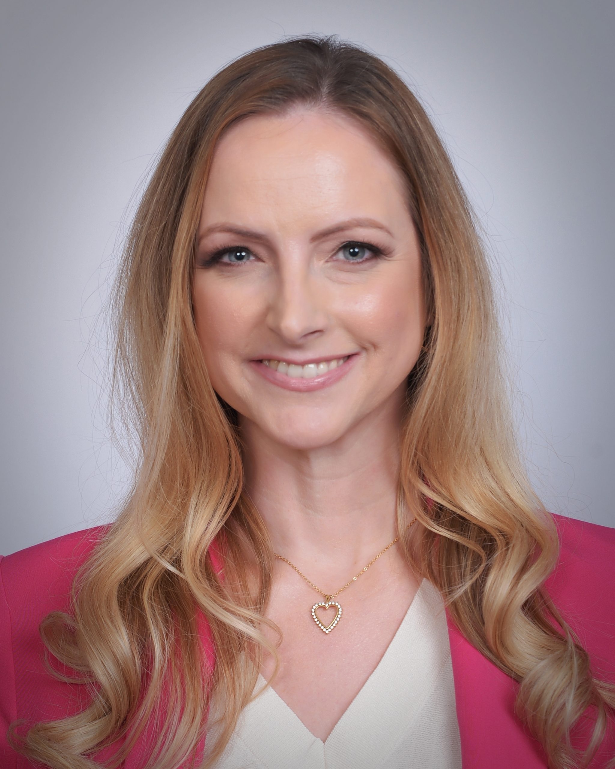 Smiling woman with long blonde hair wearing a pink blazer and heart-shaped necklace. Neutral background. Confident and friendly expression.