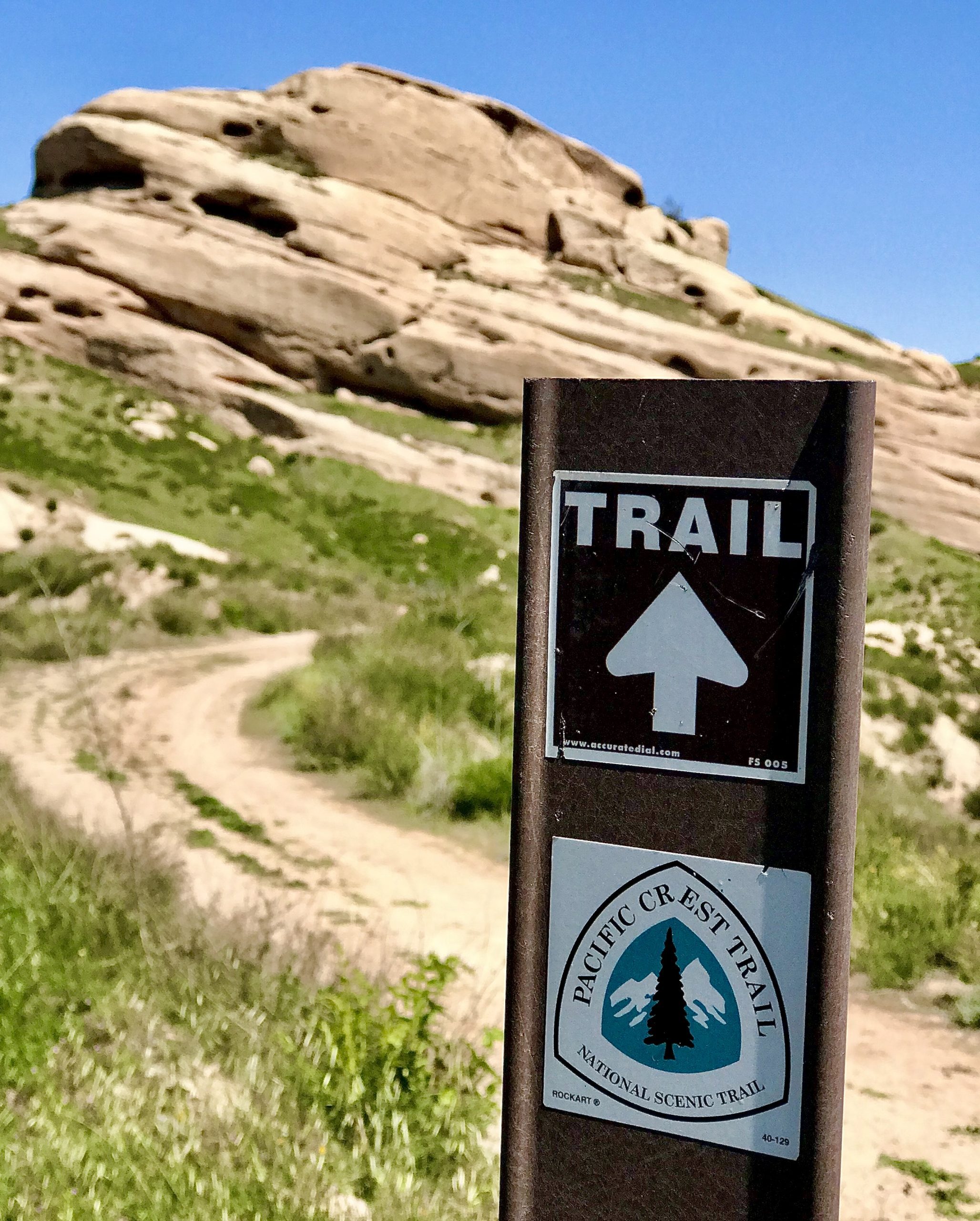 A trail sign with a white arrow and Pacific Crest Trail logo in the foreground and a winding dirt path that leads to large rock formations.