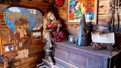 A wooden room with a vintage piano and antique doll seated on top. The walls display various items, including a cowboy-themed "Pioneertown" sign and an old movie poster.