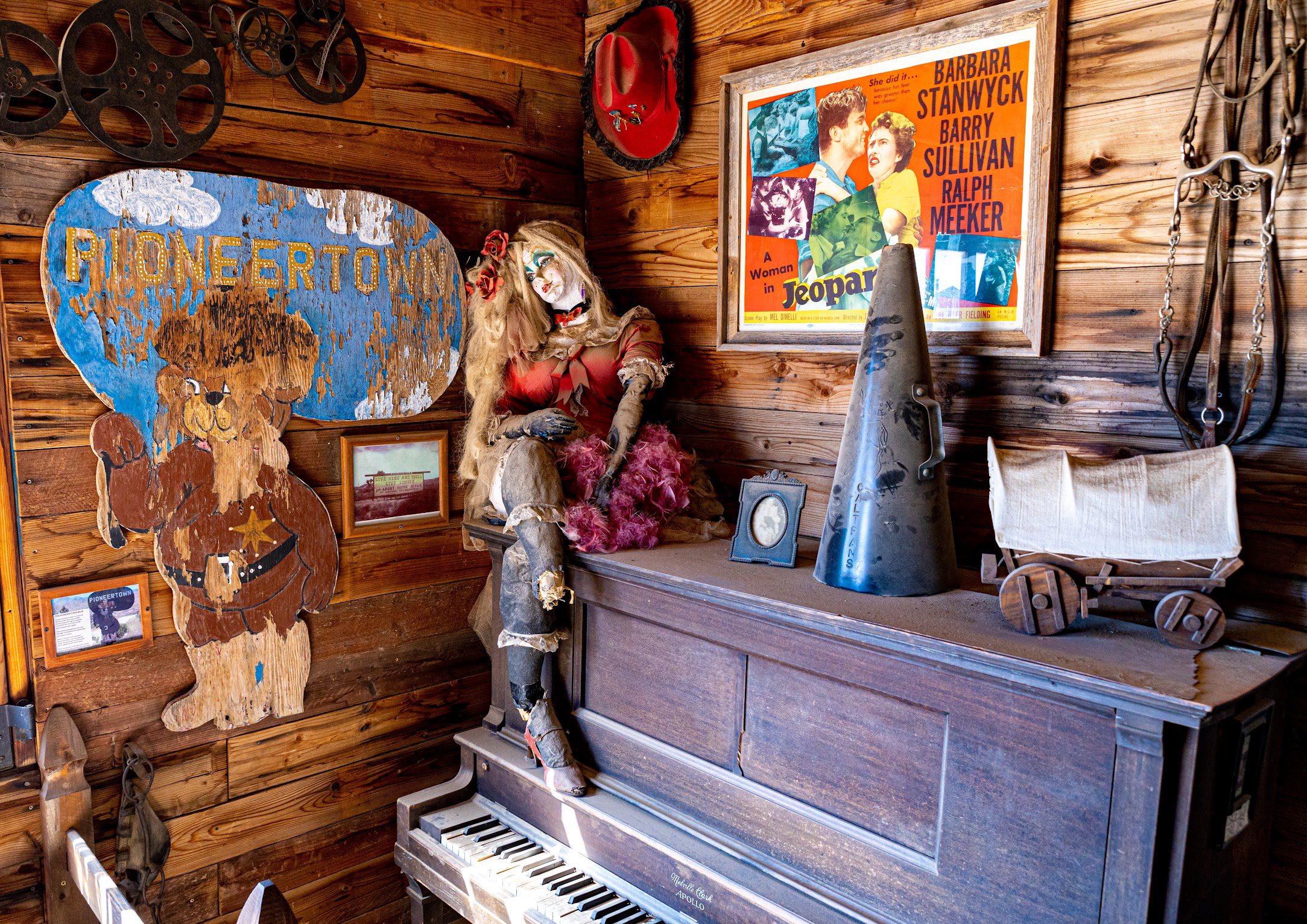 A wooden room with a vintage piano and antique doll seated on top. The walls display various items, including a cowboy-themed "Pioneertown" sign and an old movie poster.