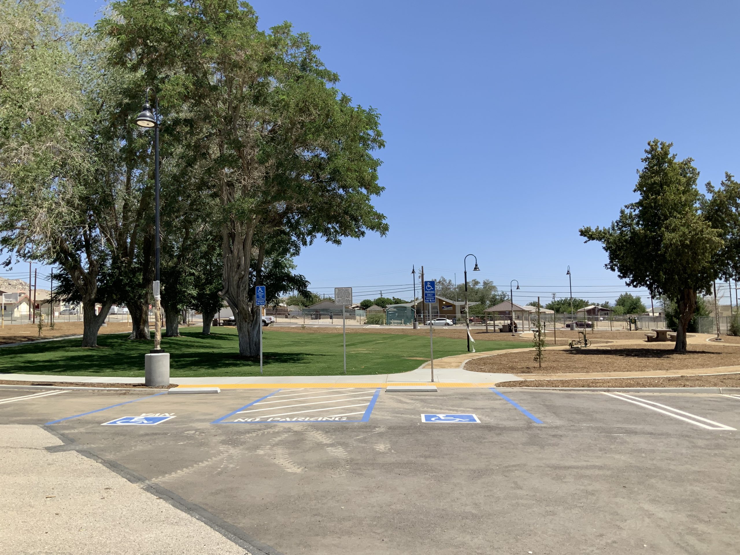 Park with trees, lawn, paths, benches, and accessible parking spaces marked with wheelchair symbols and “NO PARKING” text