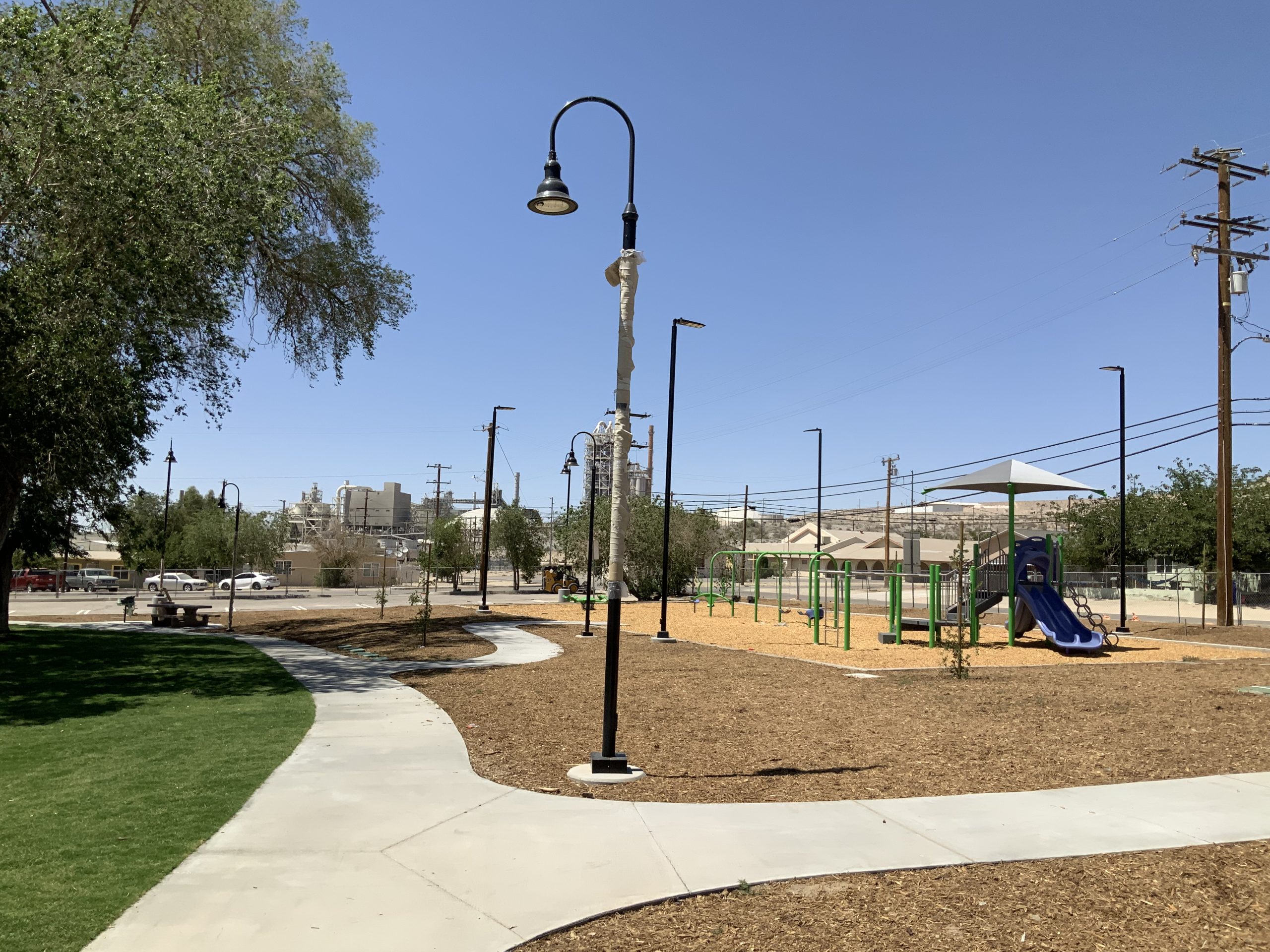 Park with curved path, trees, streetlights, and playground with slide and shade; buildings and utility poles in background.