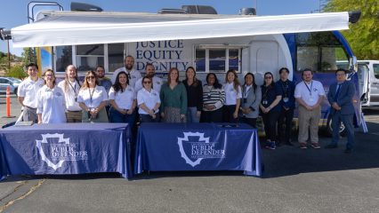 A group of people pose in front of a mobile unit labeled "Justice, Equity, Dignity" with Public Defender banners displayed on tables in front of the group.
