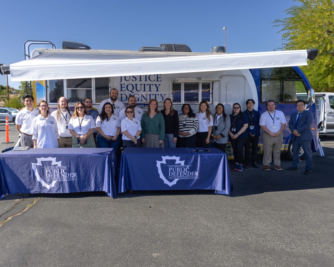 A group of people pose in front of a mobile unit labeled "Justice, Equity, Dignity" with Public Defender banners displayed on tables in front of the group.