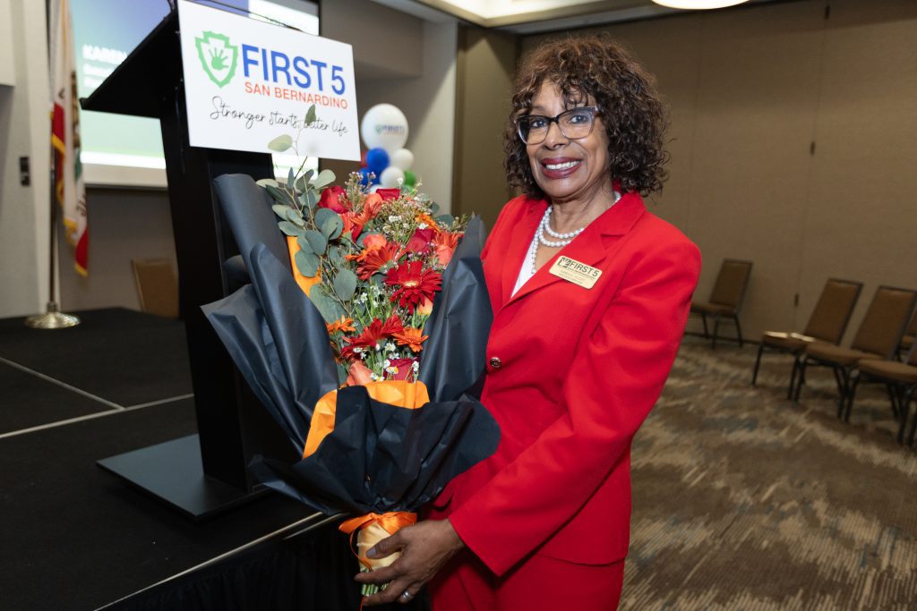 Woman in a red suit holding a bouquet, smiling beside a podium with a First 5 San Bernardino sign at an indoor event.