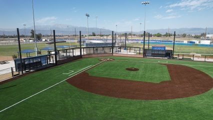 An aerial view of a newly constructed baseball field featuring synthetic turf, backstops, and protective netting, set against a scenic mountainous backdrop with additional sports facilities in the distance.