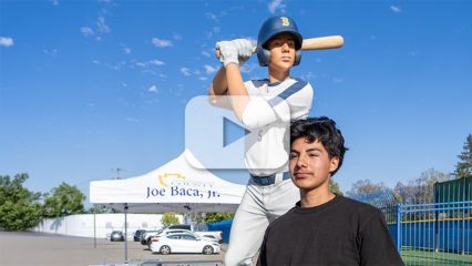 Person stands by baseball player statue; white tent reading “Joe Baca, Jr.” behind under clear blue sky, creating a bright, cheerful scene.