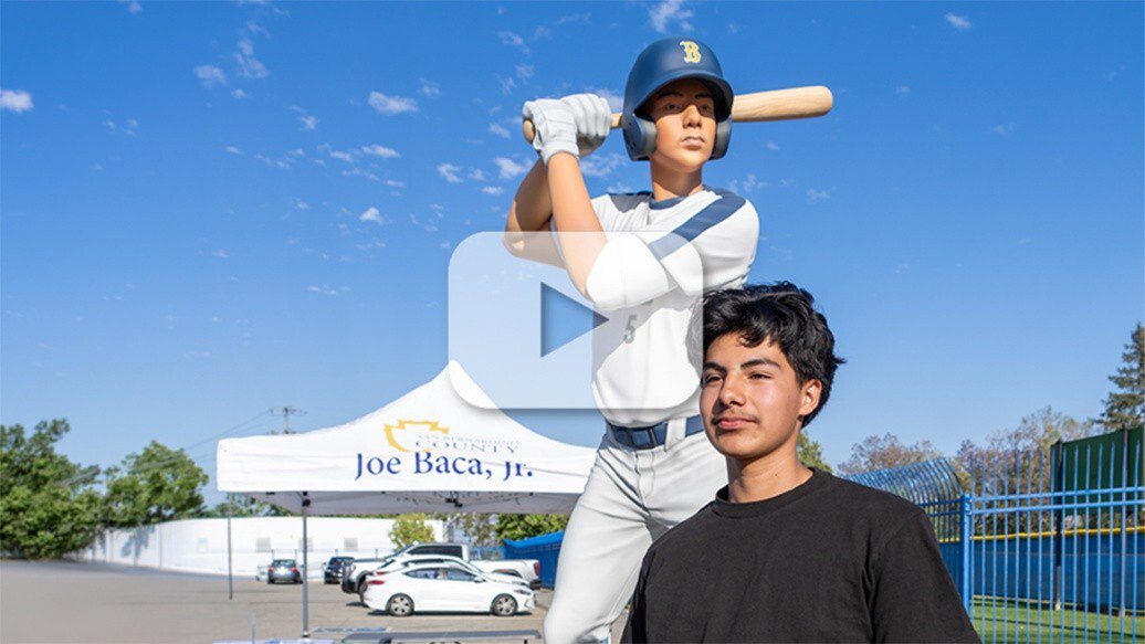 Person stands by baseball player statue; white tent reading “Joe Baca, Jr.” behind under clear blue sky, creating a bright, cheerful scene.