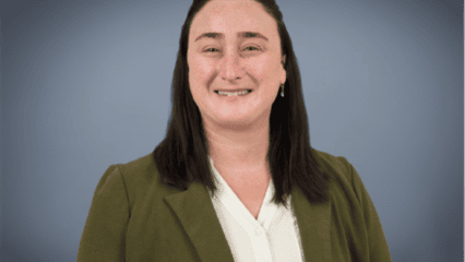 A headshot of a smiling woman wearing an olive blazer and white blouse against a soft gray background.