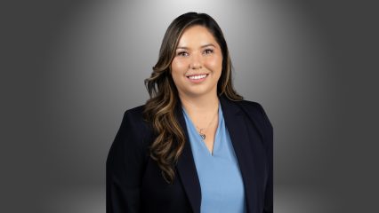 Woman with long wavy hair smiling, wearing a navy blazer and light blue blouse, posed against a grey background.