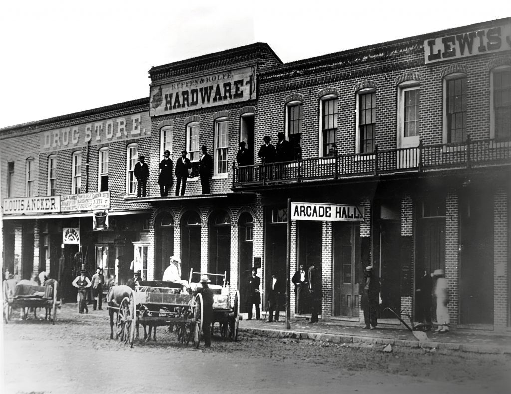 A black and white photo of buildings, including a drug store, hardware shop and an Arcade Hall. People are gathered on the sidewalks and balconies.