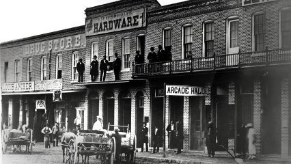 A black and white photo of buildings, including a drug store, hardware shop and an Arcade Hall. People are gathered on the sidewalks and balconies.
