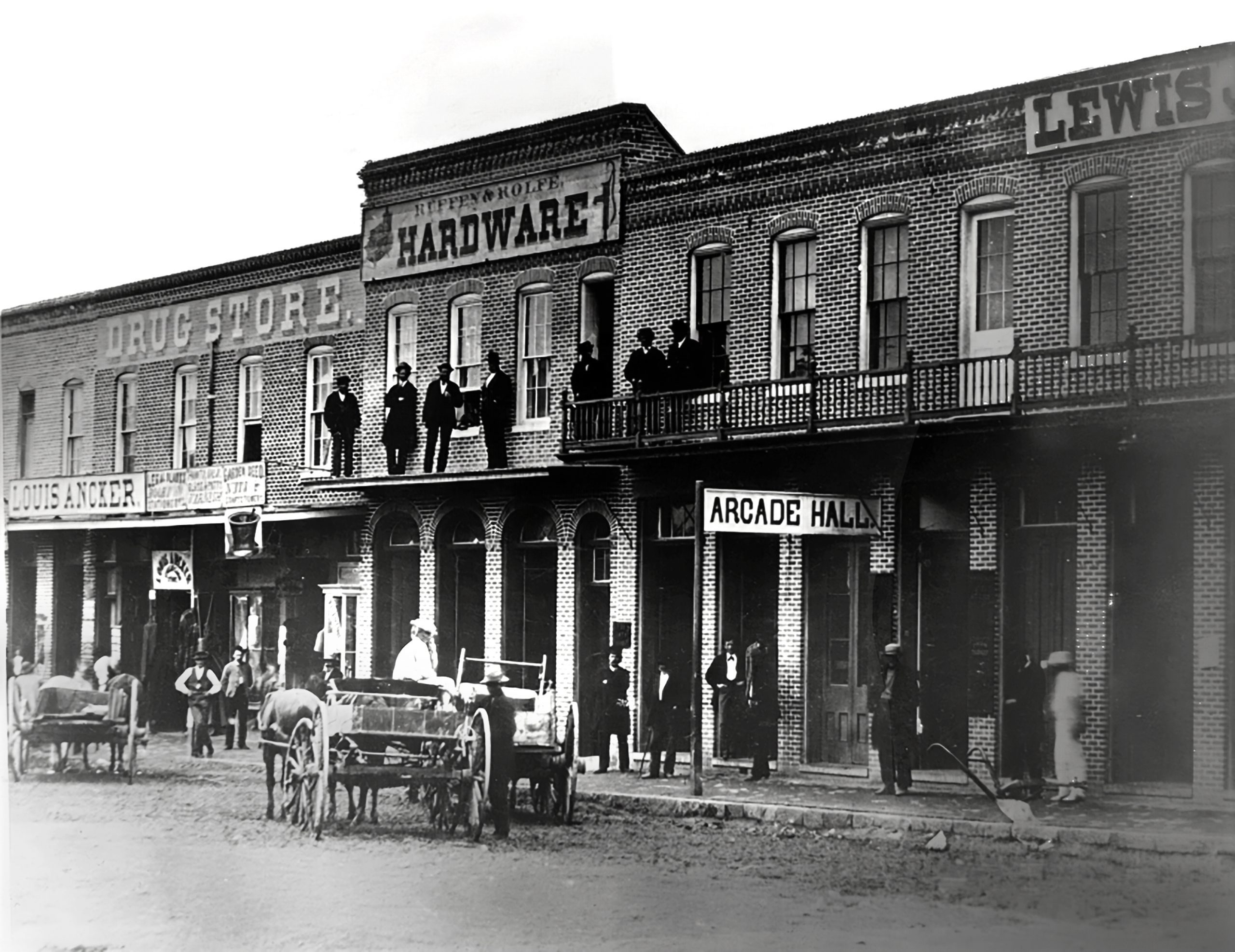 A black and white photo of buildings, including a drug store, hardware shop and an Arcade Hall. People are gathered on the sidewalks and balconies.