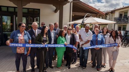A group of people stand in front of a building, gathered for a ribbon-cutting ceremony. They are holding a blue ribbon that reads "Grand Opening."