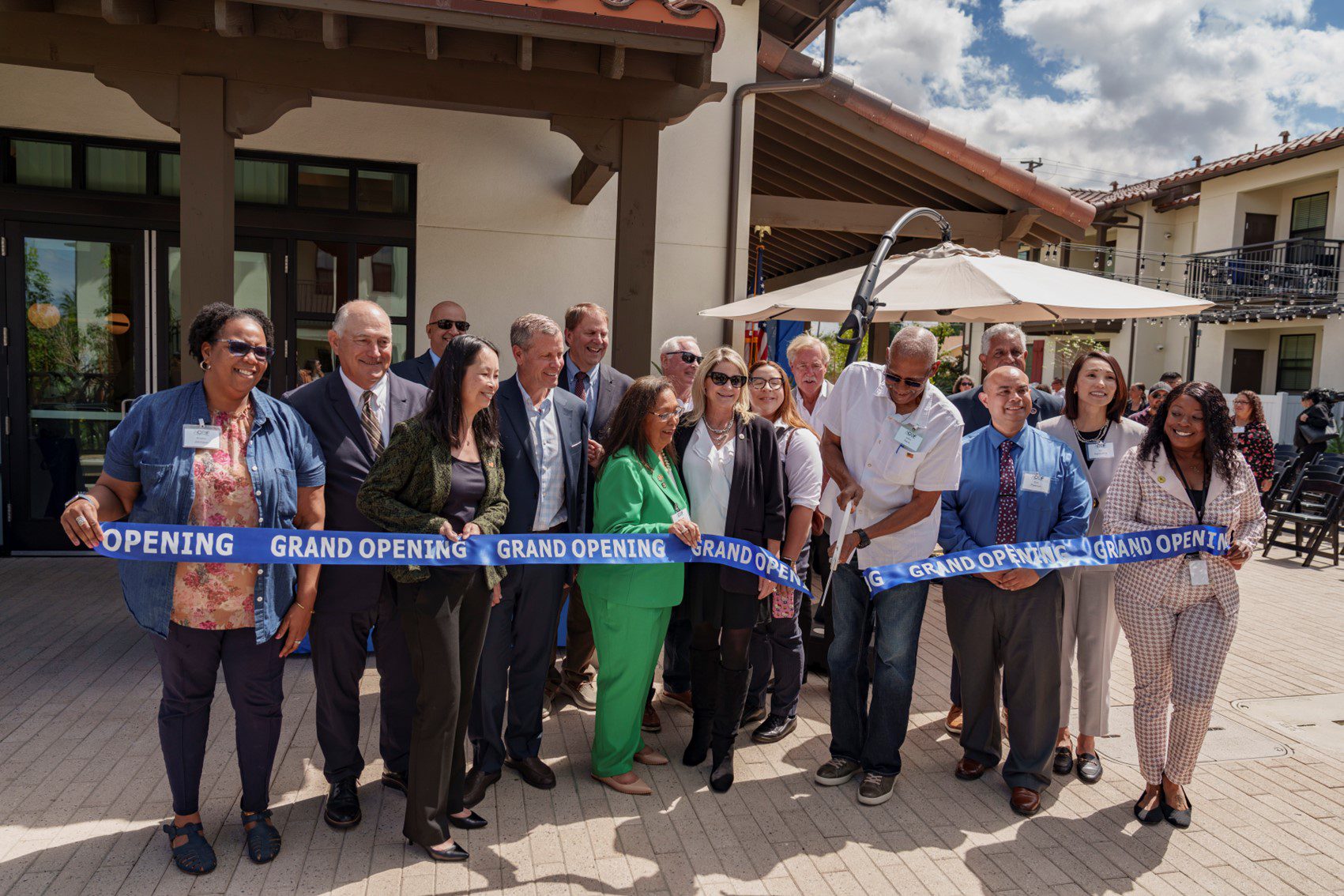 A group of people stand in front of a building, gathered for a ribbon-cutting ceremony. They are holding a blue ribbon that reads "Grand Opening."