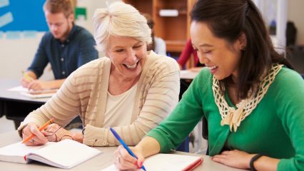 Two smiling women, one with white hair and tan cardigan, the other with black hair and a green sweater, write in notebooks at a desk with other classmates in a classroom setting.