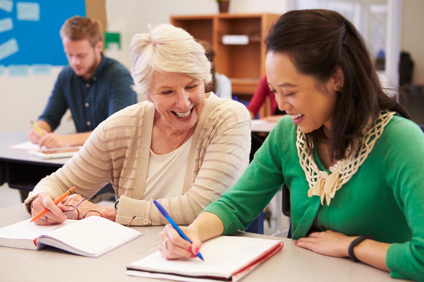 Two smiling women, one with white hair and tan cardigan, the other with black hair and a green sweater, write in notebooks at a desk with other classmates in a classroom setting.