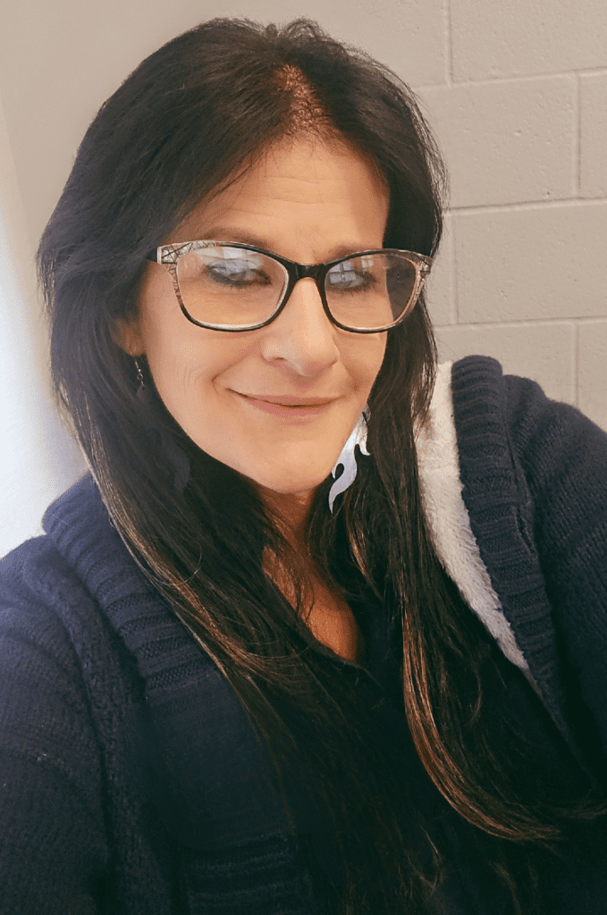 Woman with long dark hair and glasses smiling softly indoors against a light brick wall, wearing a dark sweater and earrings.