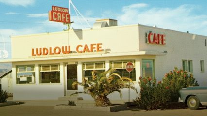 A classic diner vintage building with large windows, a red and white color scheme and a "Ludlow Cafe" sign. A vintage car is parked in front.