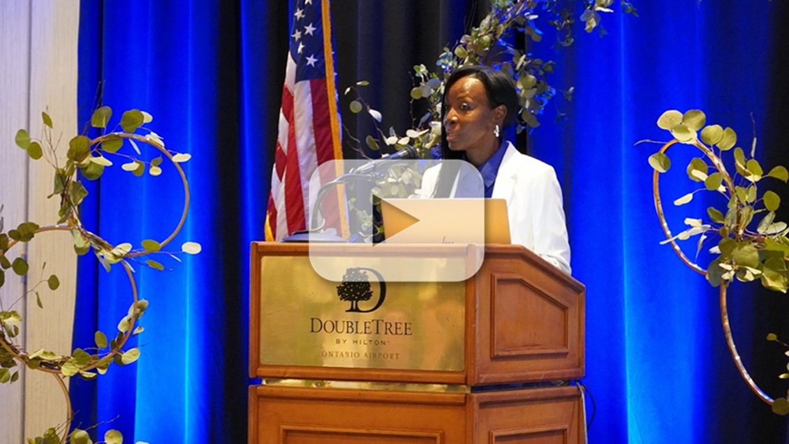 An African-American woman in a white suit with a blue shirt stands at the podium talking into a microphone with a blue back-lit curtain, a plant, and the American flag staged behind her.