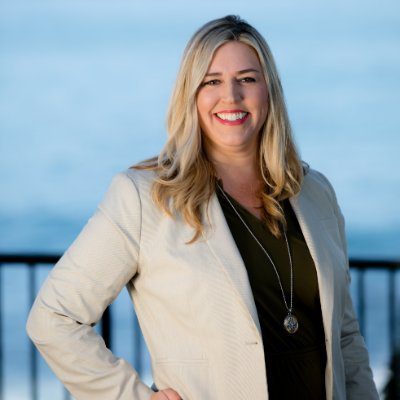 Blonde woman in a beige blazer and black top smiling outdoors by a railing with the ocean in the background.