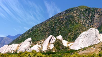 Large rock formations are in the foreground, and the background features a hillside covered with grass under a clear blue sky with wispy clouds.