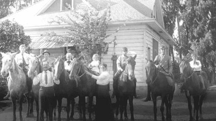 A black and white image of nine individuals posing with seven horses in front of a house.