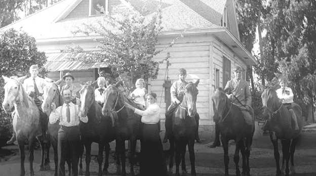 A black and white image of nine individuals posing with seven horses in front of a house.
