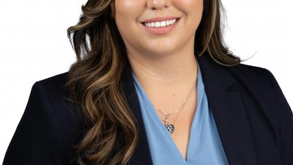 Woman with long wavy hair smiling, wearing a navy blazer and light blue blouse, posed against a white background.