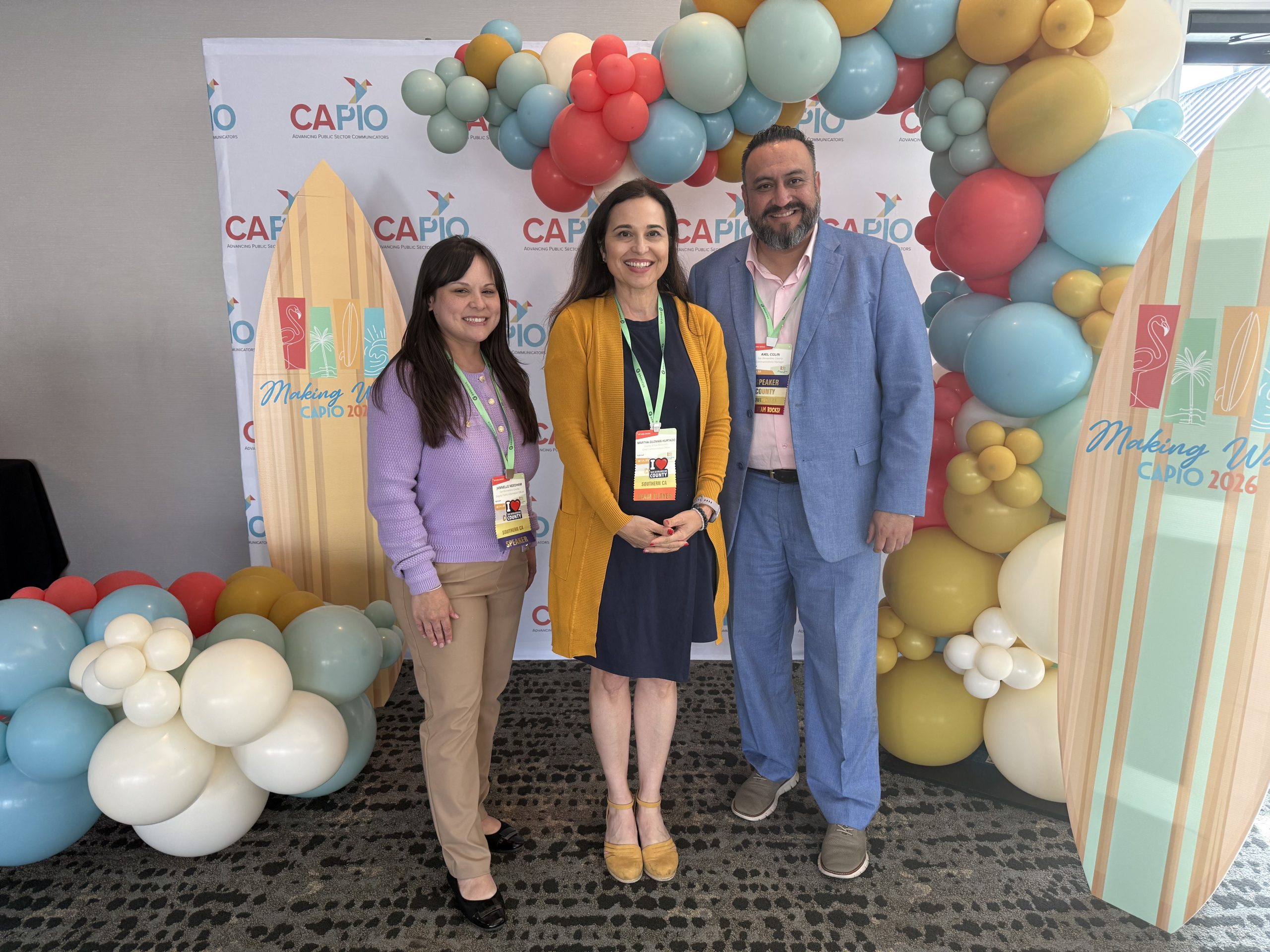 Three people pose in front of colorful balloons and a CAPIO backdrop with a “Making Waves CAPIO 2026” surfboard.