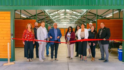 A line of people stand behind a red ribbon taped inside a horse barn while a woman in the center holds a large pair of scissors cutting the ribbon.