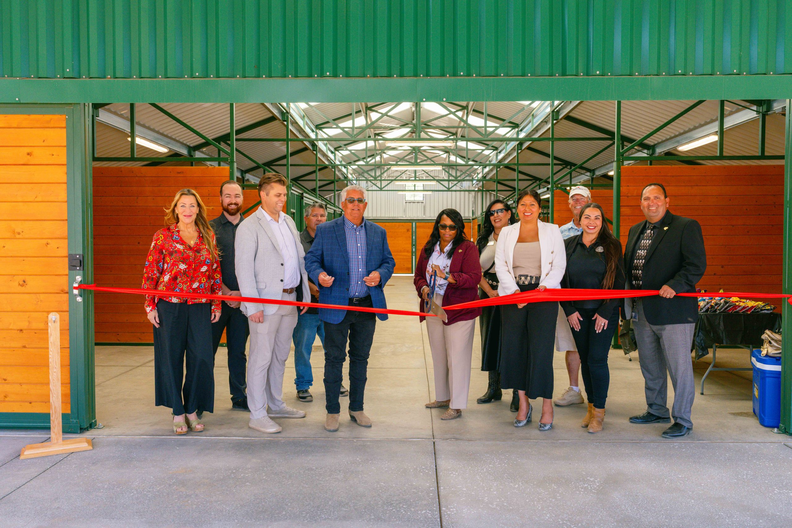 A line of people stand behind a red ribbon taped inside a horse barn while a woman in the center holds a large pair of scissors cutting the ribbon.