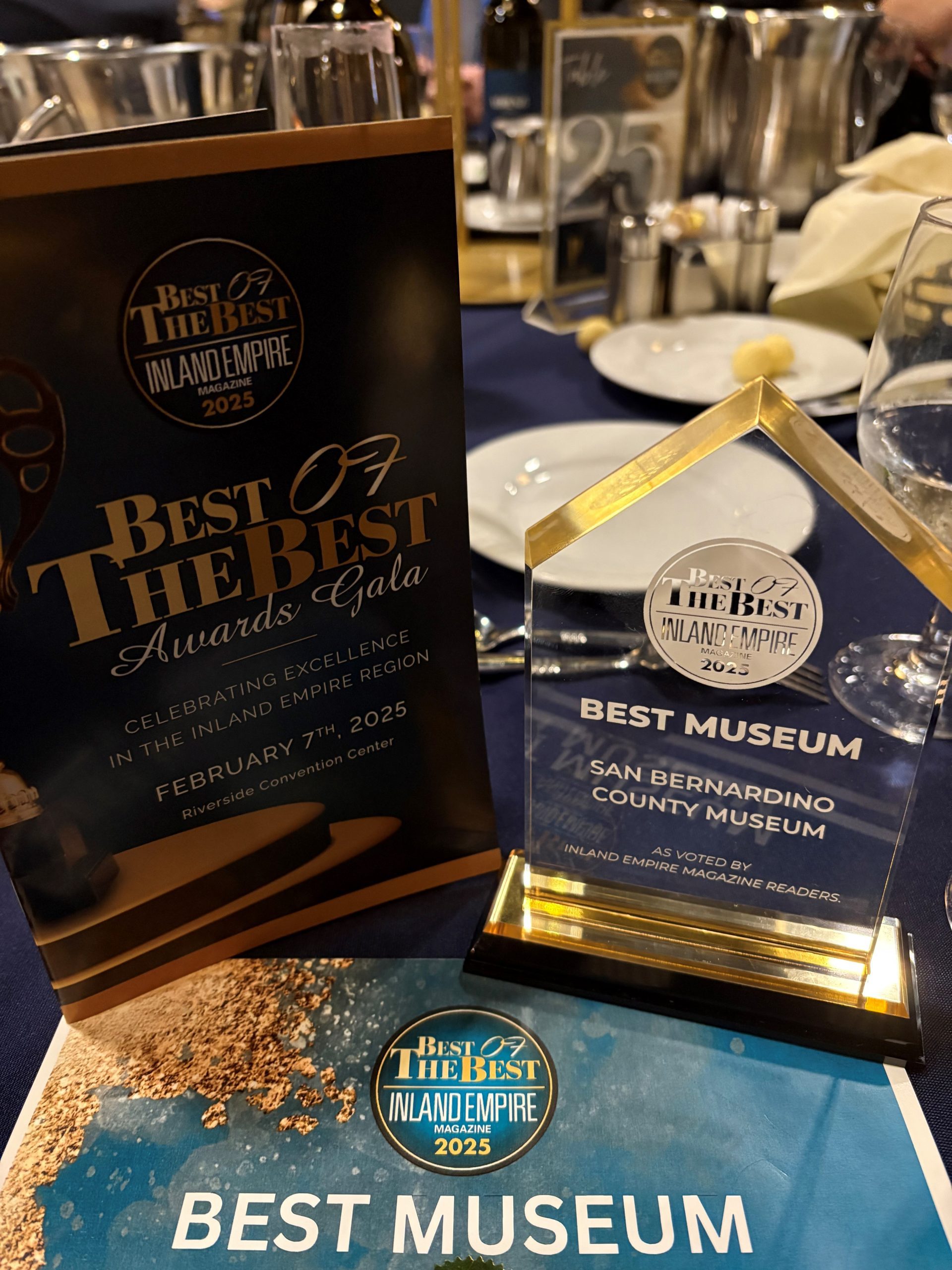 An elegant table display at the 'Best of the Best Inland Empire 2025' Awards Gala, highlighting an award presented to the San Bernardino County Museum for 'Best Museum,' with a program booklet and certificate on a navy blue tablecloth.