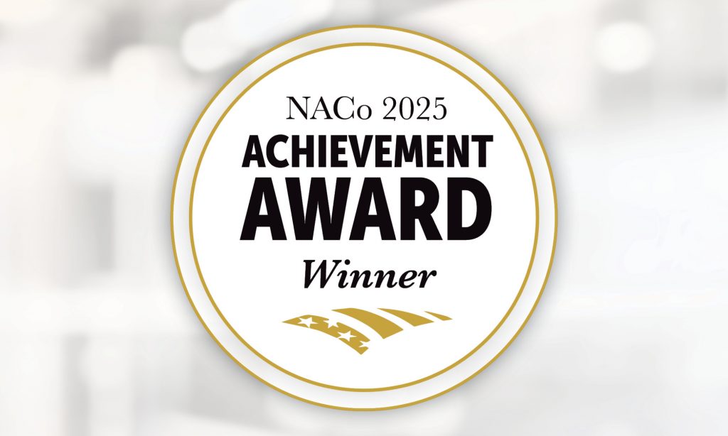 Circular NACo 2025 Achievement Award Winner badge with bold text and gold accents on a blurred white background.