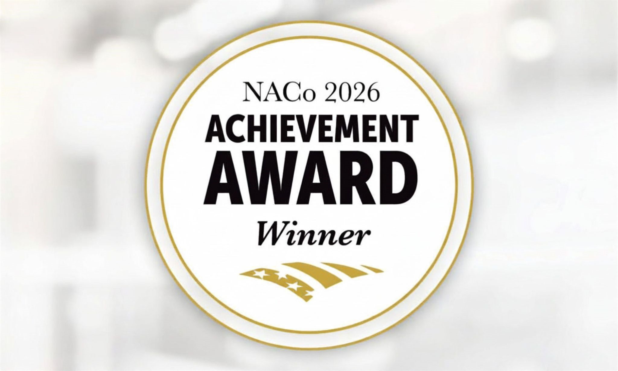 Circular gold-and-white seal with the text “NACo 2026 Achievement Award Winner” against a soft blurred background.