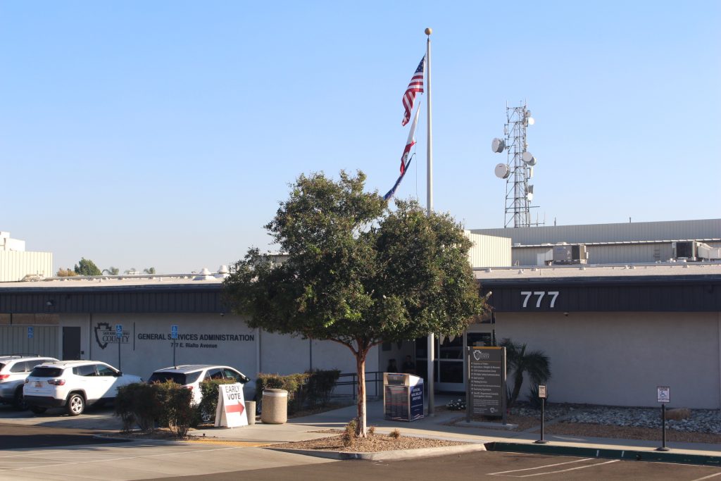 Exterior of a General Services Administration building with a U.S. flag, parked cars, and a sign directing voters to early voting.