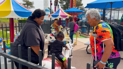 Two people stand on either side of a toddler on a ride at an amusement park.